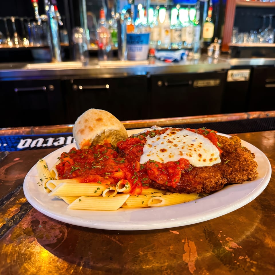 Chicken Parmesan with marinara, melted cheese, penne pasta, and a bread roll served at a warmly lit bar.