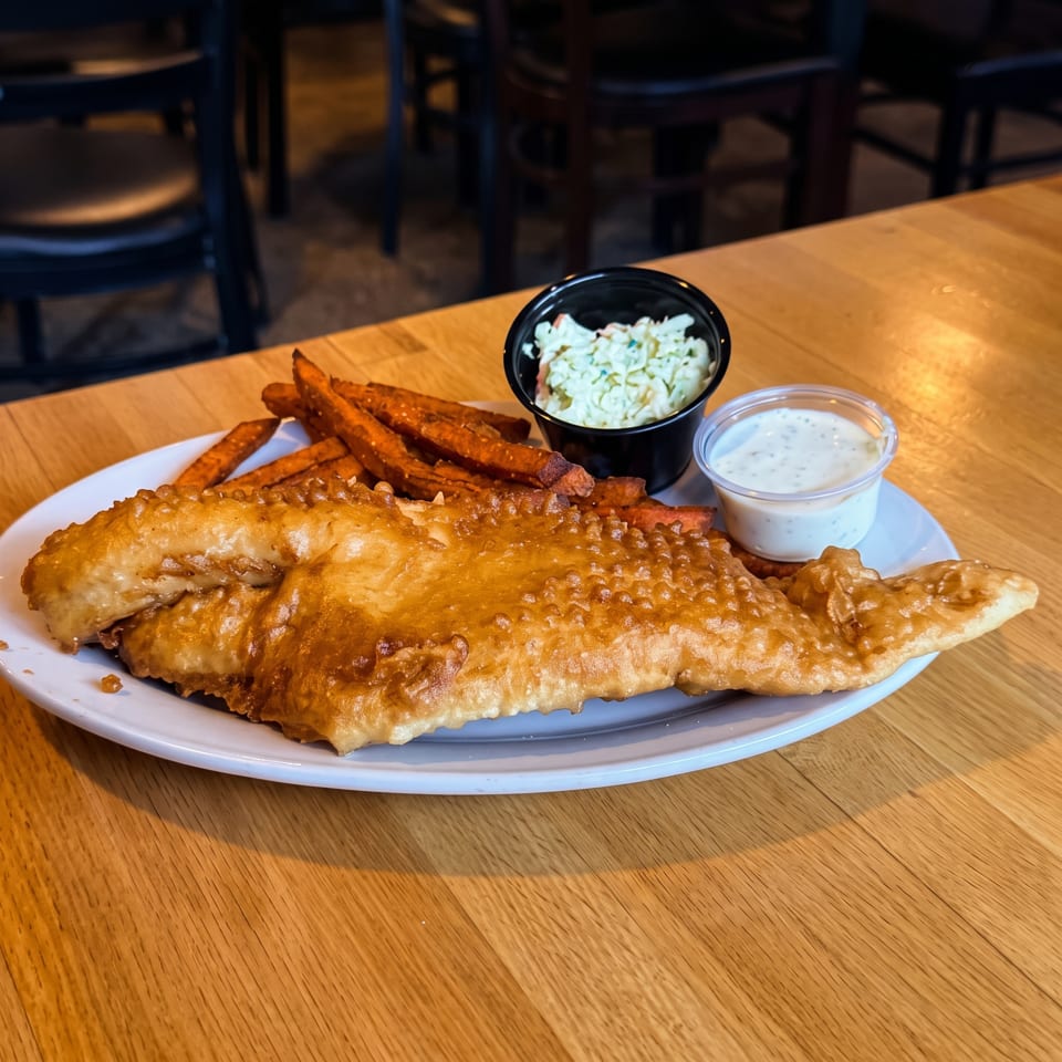 Golden-battered fish with sweet potato fries, coleslaw, and tartar sauce served on a white plate in a restaurant.