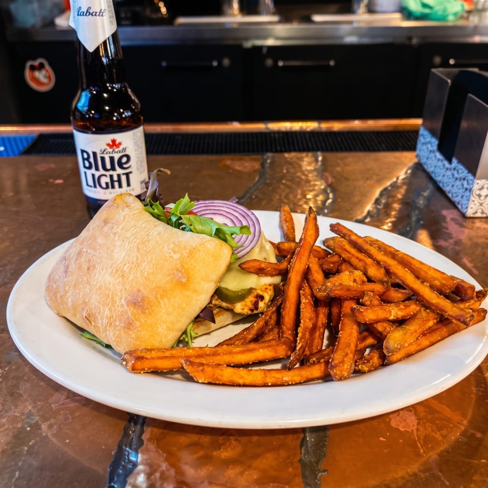 Grilled sandwich with lettuce, onions, pickles, and cheese served with sweet potato fries beside a Labatt Blue Light beer.